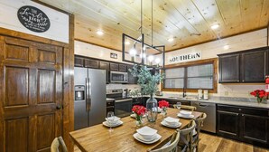 A kitchen with wooden cabinets, stainless steel appliances, and a dining table set for four. - Private Indoor Pool Cabin | Arcade, Hot Tub | 1.5mi to Parkway (Pigeon Forge)