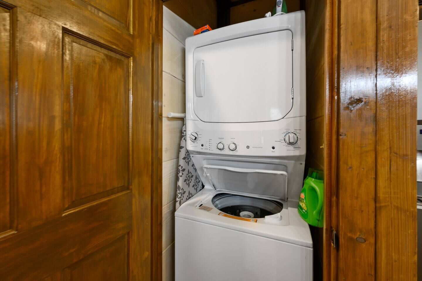 A compact washer and dryer unit in a wooden cabinet with a green detergent bottle beside it.