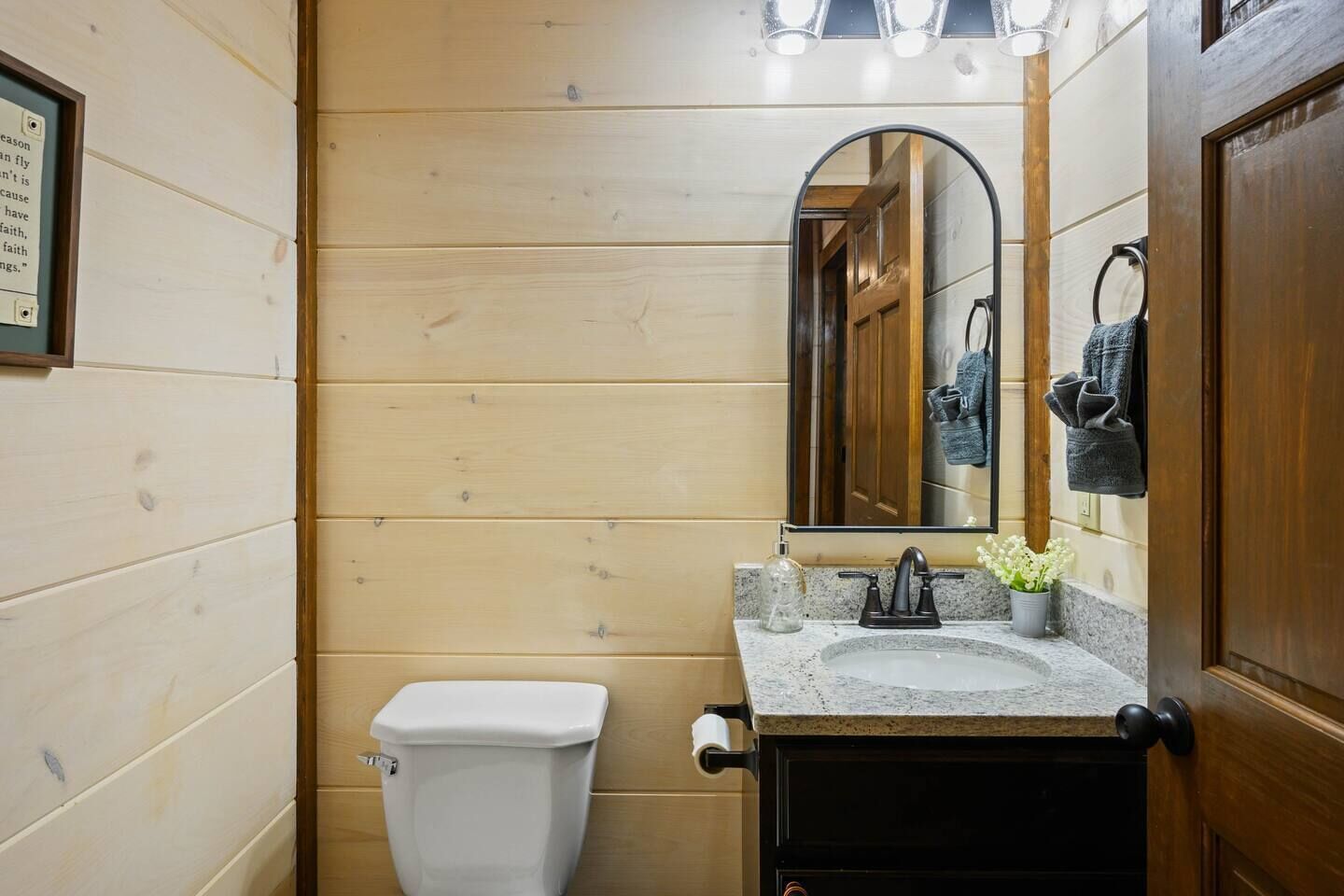 A compact bathroom with a wooden wall, a white toilet, a granite sink, a mirror, and a towel rack.