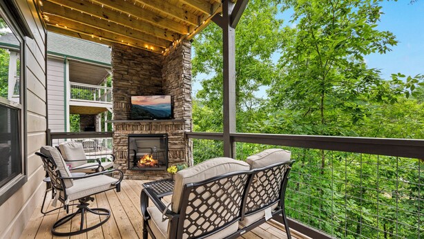 A covered deck with a stone fireplace, outdoor seating, and a view of a lush green landscape. - Private Indoor Pool Cabin | Arcade, Hot Tub | 1.5mi to Parkway (Pigeon Forge)
