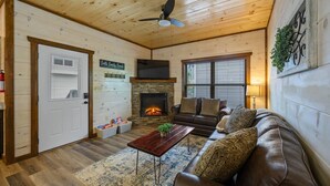 A cozy cabin interior with a stone fireplace, wooden walls, a leather sofa, and a dining table. - Private Indoor Pool Cabin | Arcade, Hot Tub | 1.5mi to Parkway (Pigeon Forge)