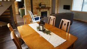 Een houten eettafel met een witte placemat en een lantaarn, omringd door houten stoelen. Achtergrond bevat een open haard en een aan de muur bevestigde televisie.