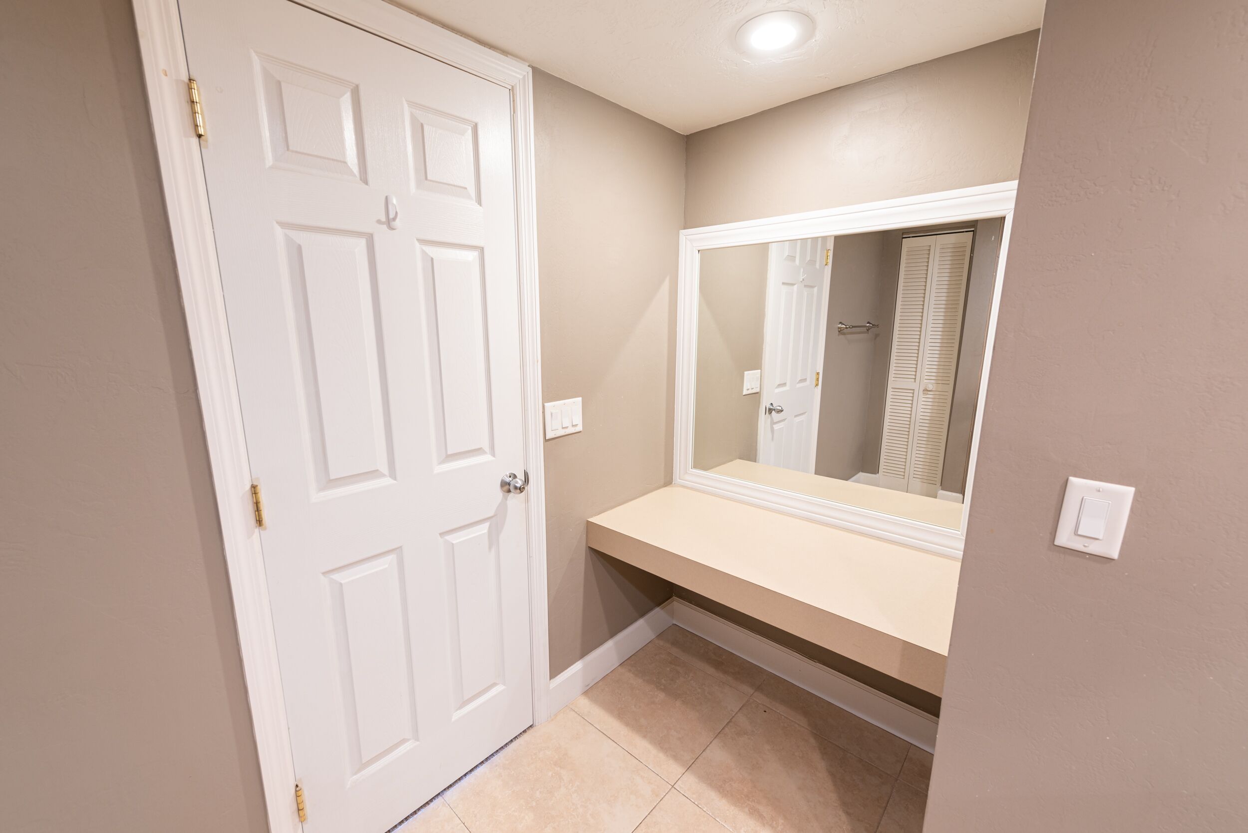 A bathroom with a white door, beige walls, and a mirror above a countertop.