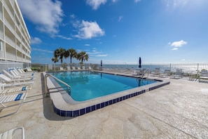 A pool with lounge chairs and palm trees, a clear sky, and a view of the ocean.