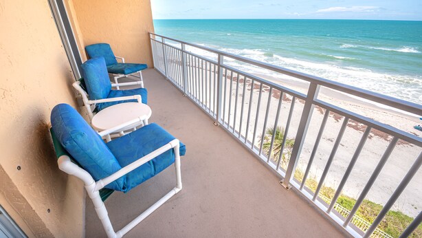 Ein Balkon mit zwei blauen Sitzgelegenheiten und einem kleinen Tisch, mit Blick auf einen Strand, Wellen und einen klaren Himmel.