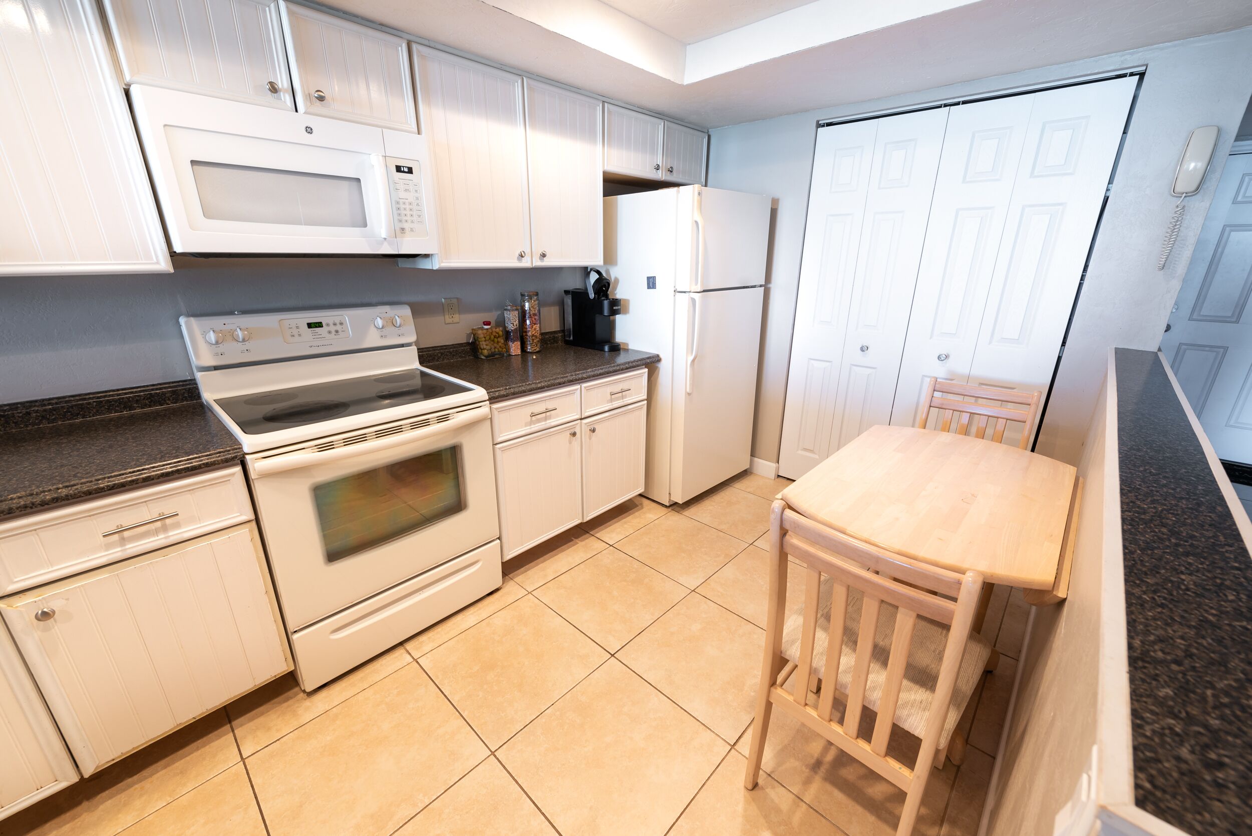 A kitchen with white cabinets, a microwave, a stove, a refrigerator, and a wooden table with a chair.