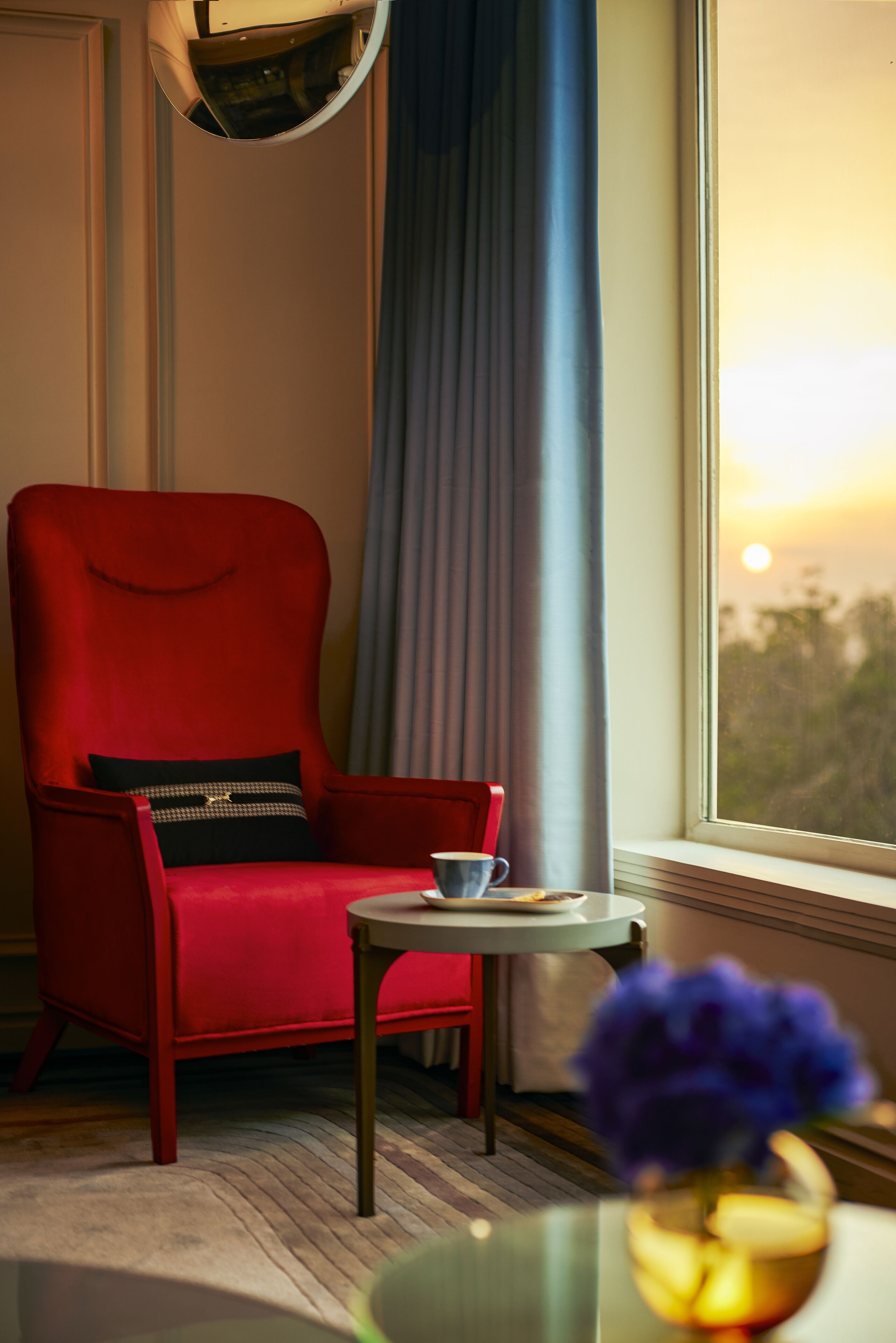 A red armchair with a black cushion, a small round table with a cup, and a vase with flowers by a window with curtains.