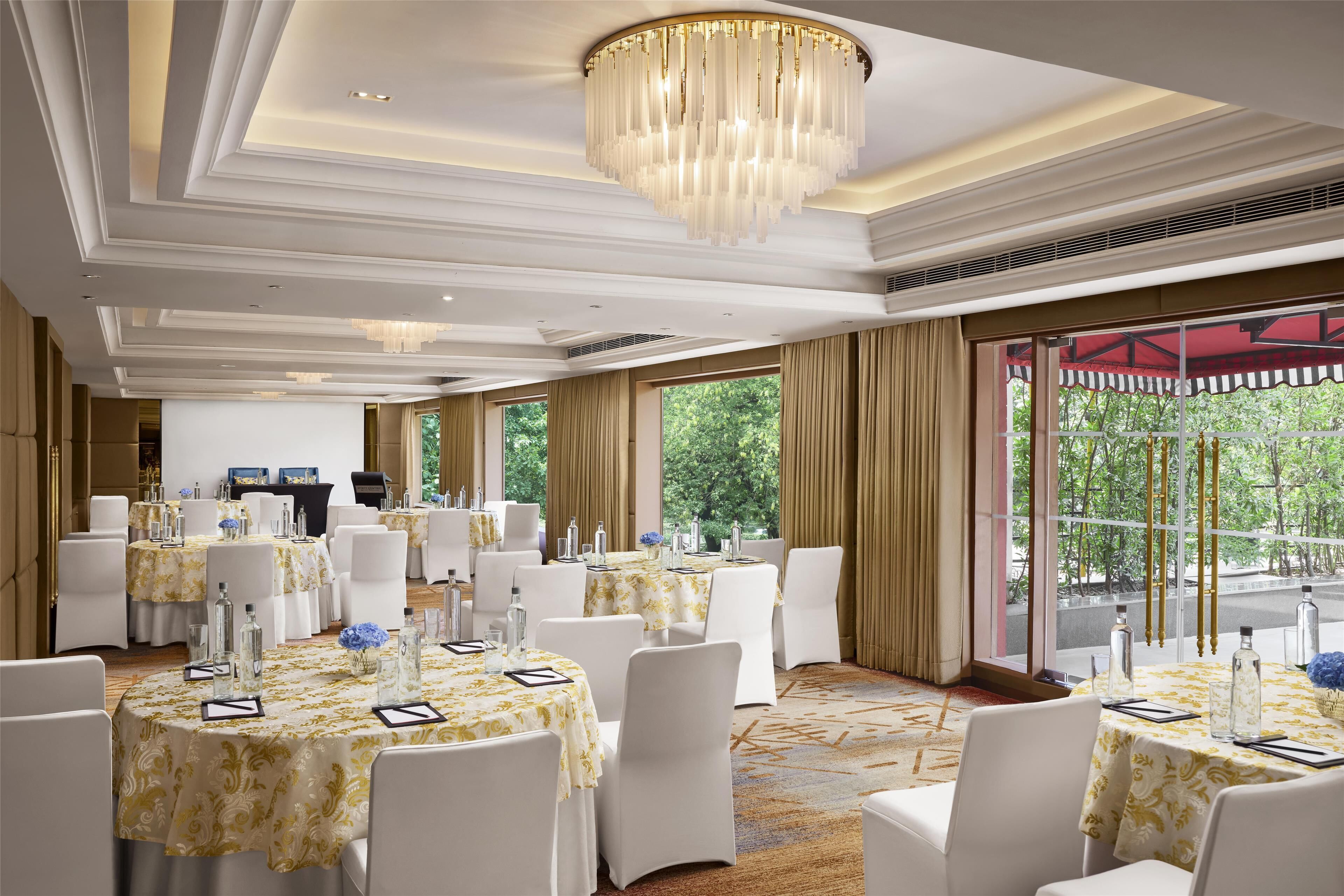 A banquet hall with round tables, white chairs, chandeliers, and large windows with curtains.