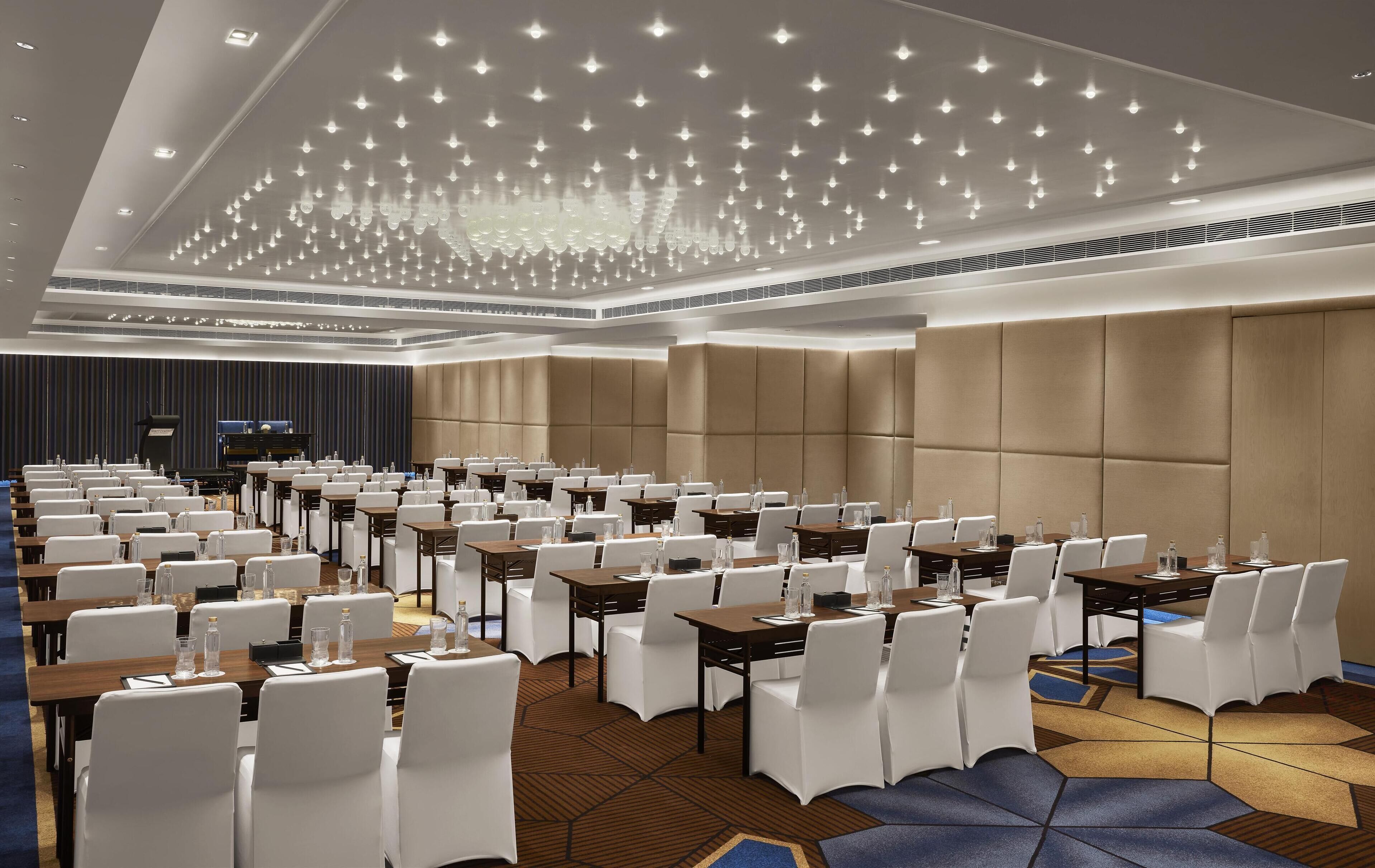 A conference room with rows of tables and chairs, a stage, and a modern chandelier.