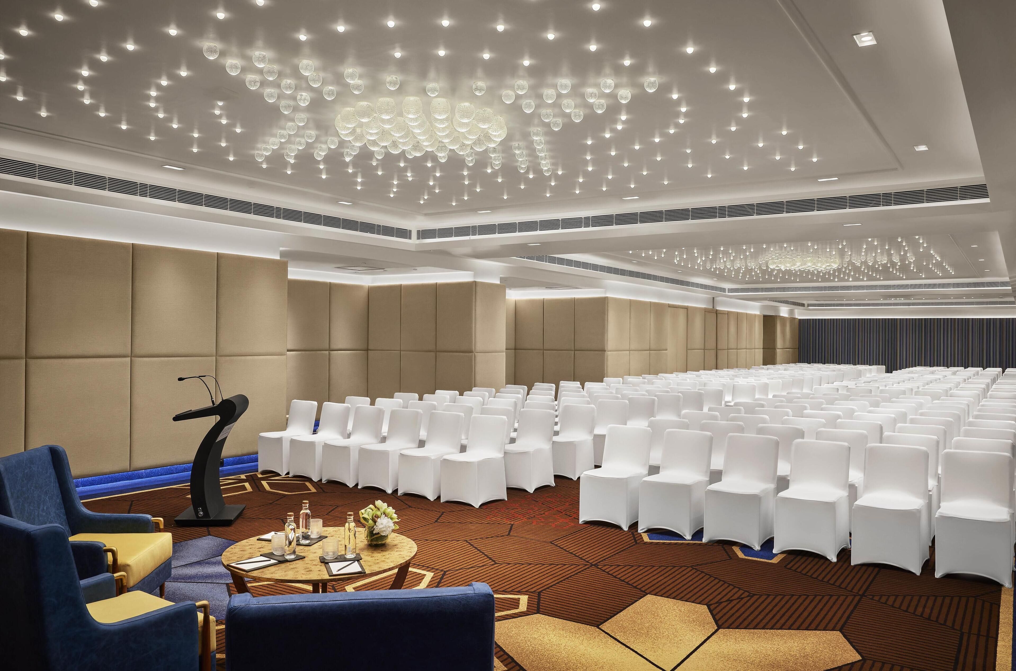 A conference room with rows of white chairs, a podium, and a modern chandelier.