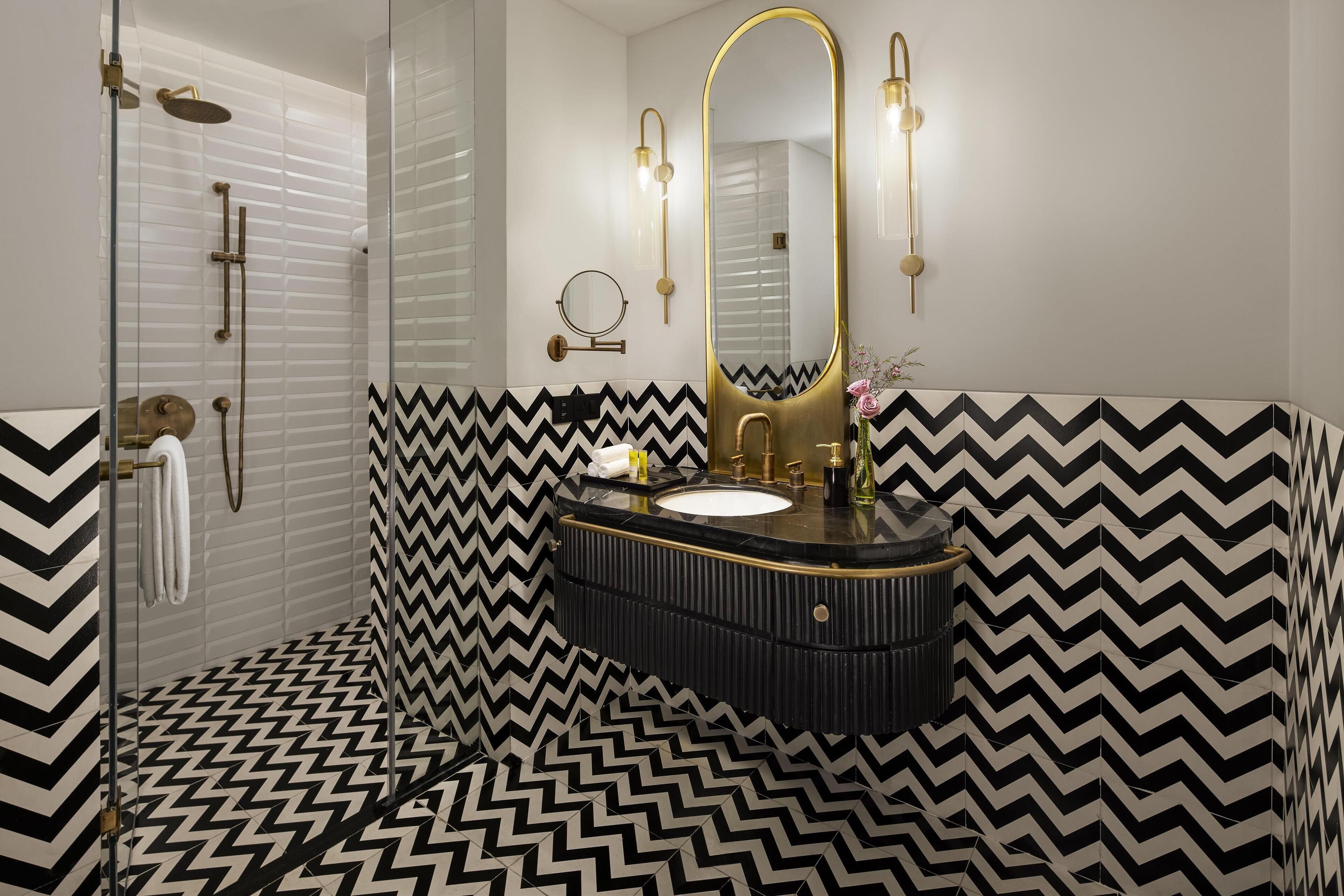 Un baño moderno con un patrón de baldosas a cuadros blancos y negros, un espejo con marco dorado y una zona de ducha con mampara de vidrio.