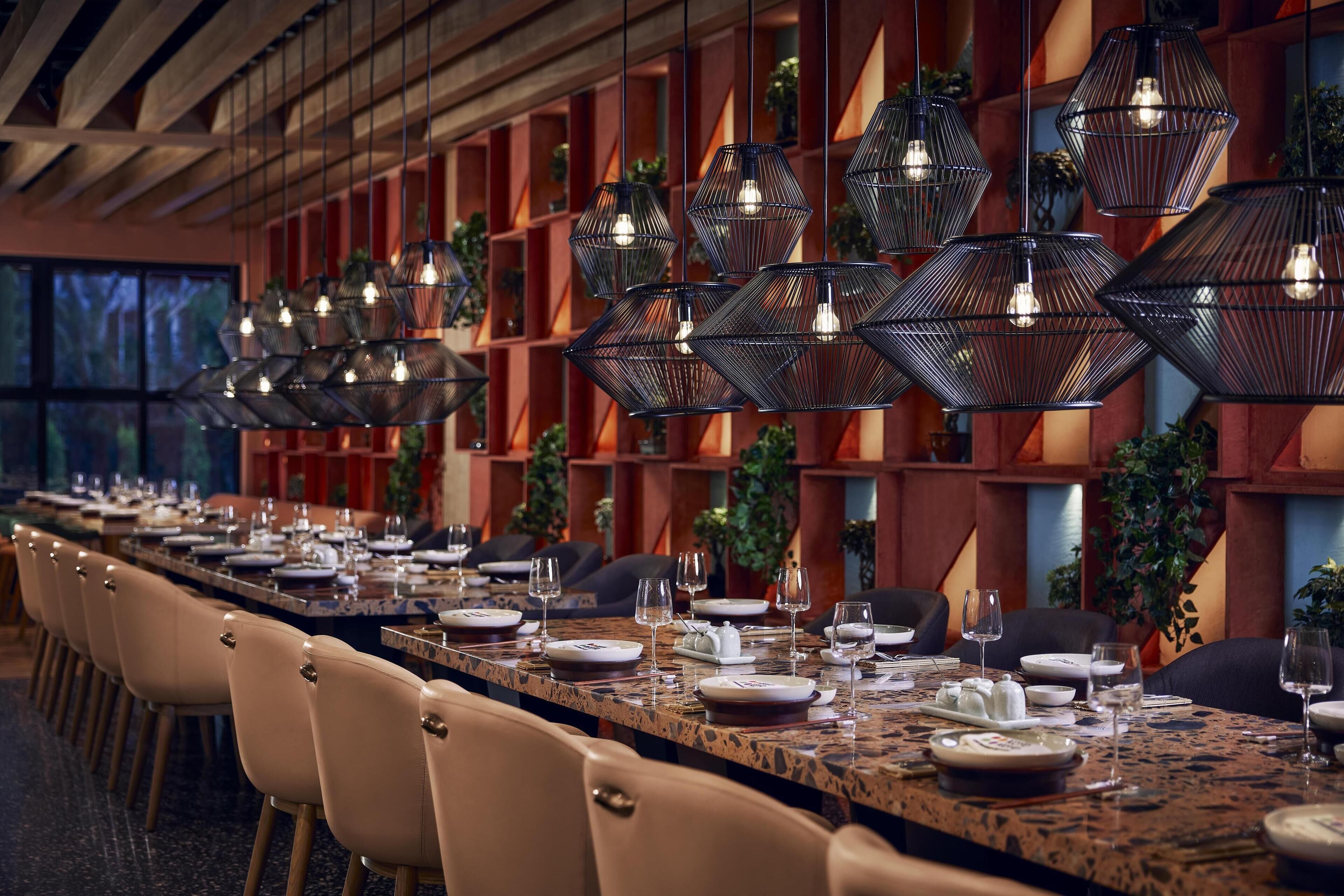 A long dining table set with chairs, surrounded by geometric wall panels and hanging pendant lights.