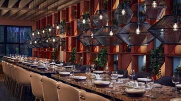 A long dining table set with chairs, surrounded by geometric wall panels and hanging pendant lights.