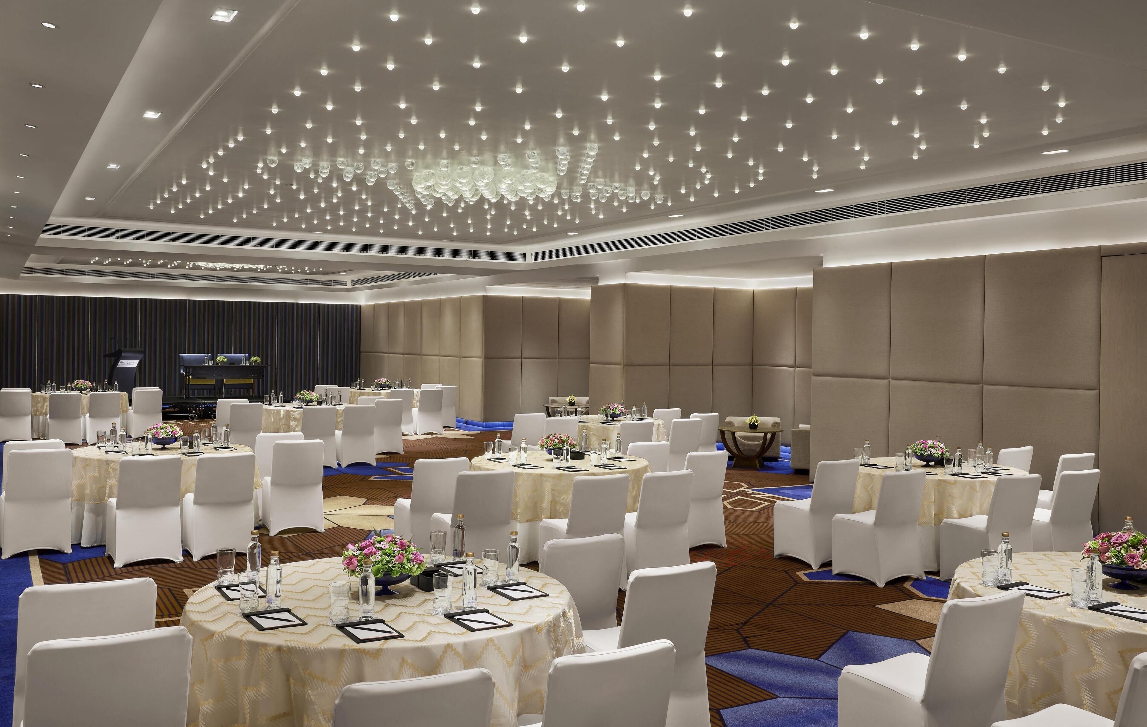 A conference room with round tables, white chairs, and a ceiling with hanging lights.