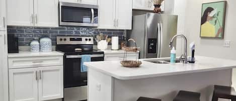 A modern kitchen with white cabinets, stainless steel appliances, a central island with stools, and a ceiling fan.