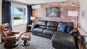 Ein modernes Wohnzimmer mit einer Ledersofa, einem Ledersessel und einem kleinen Hocker. Ein Fenster mit Vorhängen, ein Deckenventilator und eine Ziegelwand mit einem Bild sind zu sehen.