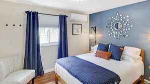 Ein Schlafzimmer mit einem Bett, einem Sessel, einem Fenster mit Vorhängen, einer Klimaanlage, einem Wandspiegel und einer Wandlampe.