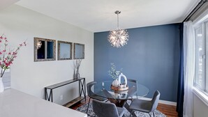 Ein moderner Essbereich mit einem Glastisch, grauen Stühlen und einer Chandelier. An der Wand befinden sich drei gerahmte Spiegel und ein Beistelltisch mit Blumen.