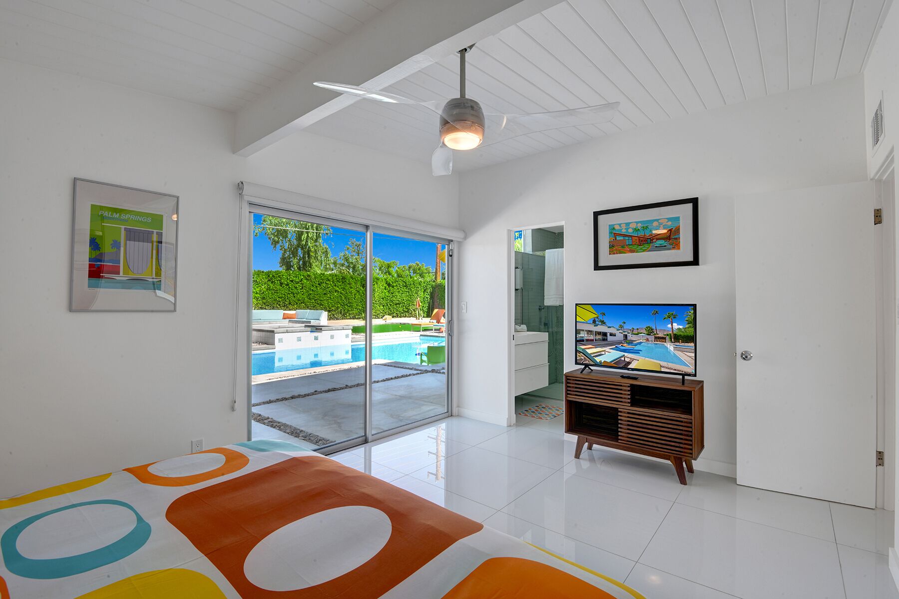 A modern living room with a large TV, a ceiling fan, and a view of a swimming pool through glass doors.