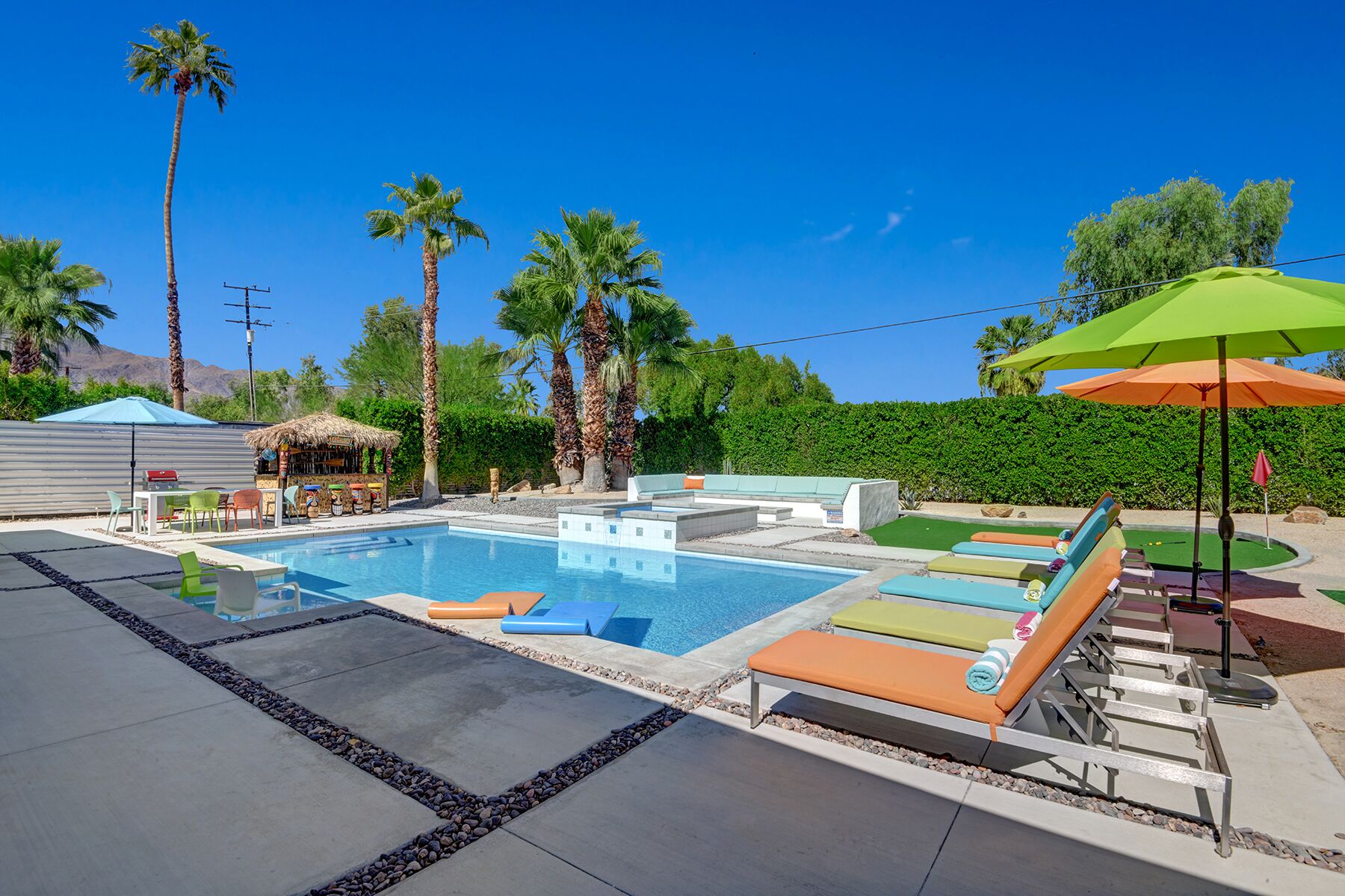 A swimming pool with lounge chairs and umbrellas, surrounded by palm trees and a barbecue area.