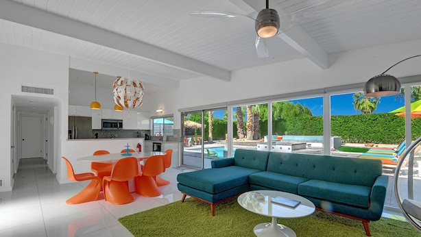 Un salon moderne avec un canapé d’angle, un coin repas avec des chaises orange, une cuisine avec des meubles blancs et une grande fenêtre donnant sur une piscine et un espace verdoyant.