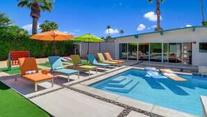 Un espace piscine moderne, doté de chaises longues et de parasols, entouré de palmiers et surplombé d’un ciel bleu azur.