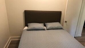 Ein Bett mit dunklem Kopfteil und zwei Kissen, eine weiße Wand und eine Tür rechts.