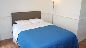 Un letto rifatto con una coperta blu e cuscini bianchi, una lampada da terra alta e un pavimento in legno.