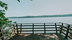 A wooden deck with a railing overlooking a calm lake. - The Chalet is located in the woods of a gated property with a covered porch. (Darlington)