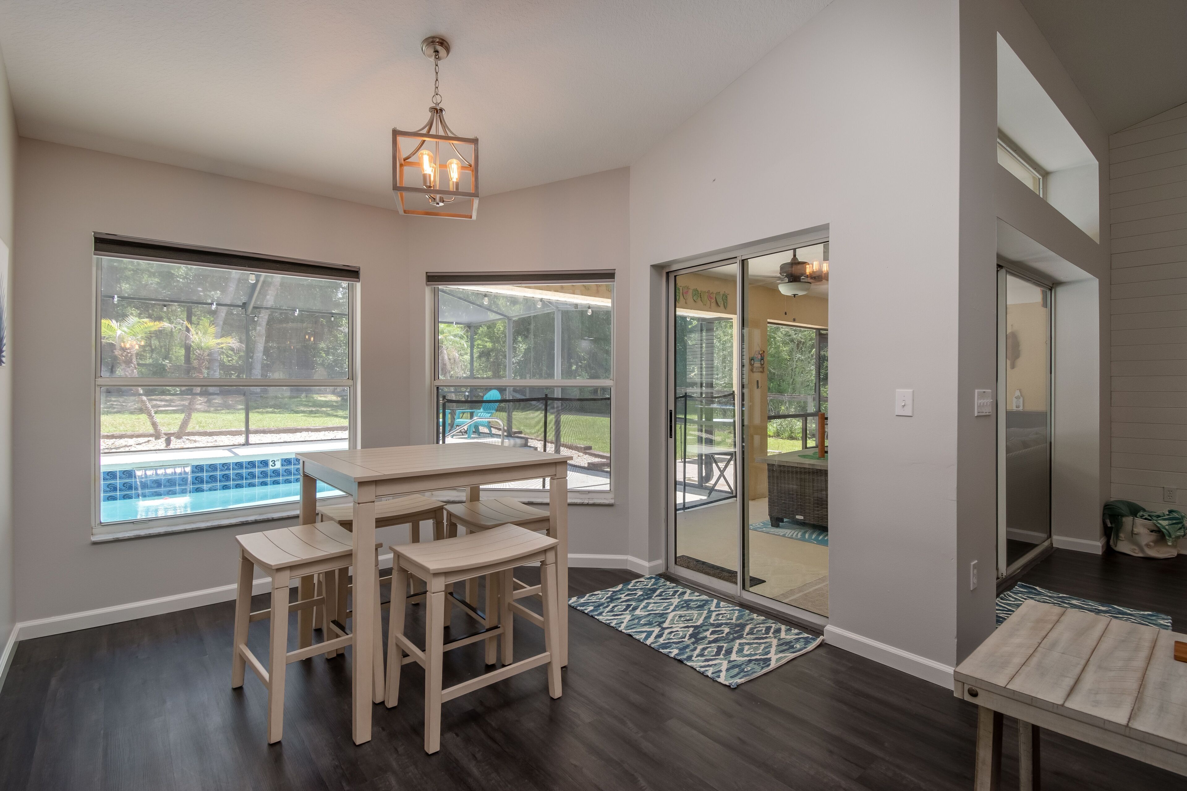 A modern kitchen with dark wood flooring, a wooden dining table with chairs, and large windows overlooking a backyard with a pool.
