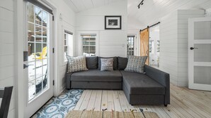 A modern living room with a gray sectional sofa, patterned cushions, a framed picture on the wall, and a large window. - The Cambridge Cove! Hot tub & Outdoor Gas Firepit (Fort Erie)