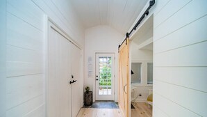 A narrow hallway with white wooden walls and a wooden floor. There is a door with a window at the end, a small plant, and a desk with a chair. - The Cambridge Cove! Hot tub & Outdoor Gas Firepit (Fort Erie)