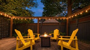 An outdoor deck with wooden flooring, a central fire feature, and yellow Adirondack chairs. - The Cambridge Cove! Hot tub & Outdoor Gas Firepit (Fort Erie)