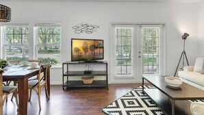 A dining area with a wooden table and chairs, a television on a stand, and a sofa with a floor lamp.