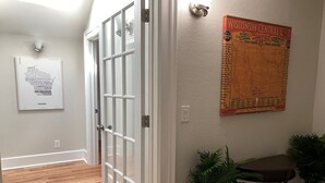Un couloir avec un plancher en bois, une porte blanche dotée de panneaux de verre, une carte accrochée au mur et deux plantes en pot.