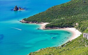 A coastal landscape with a beach, clear turquoise waters, and a forested hill. - Sportive Life - Setubal (Casal das Figeiras)