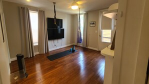 Een ruime woonkamer met houten vloeren, een wandgemonteerde televisie en fitnessapparatuur.