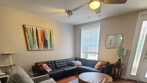 Een woonkamer met een hoekbank, een grijze fauteuil, een ronde salontafel, een plafondventilator, een kleurrijk abstract schilderij en een raam met jaloezieën.