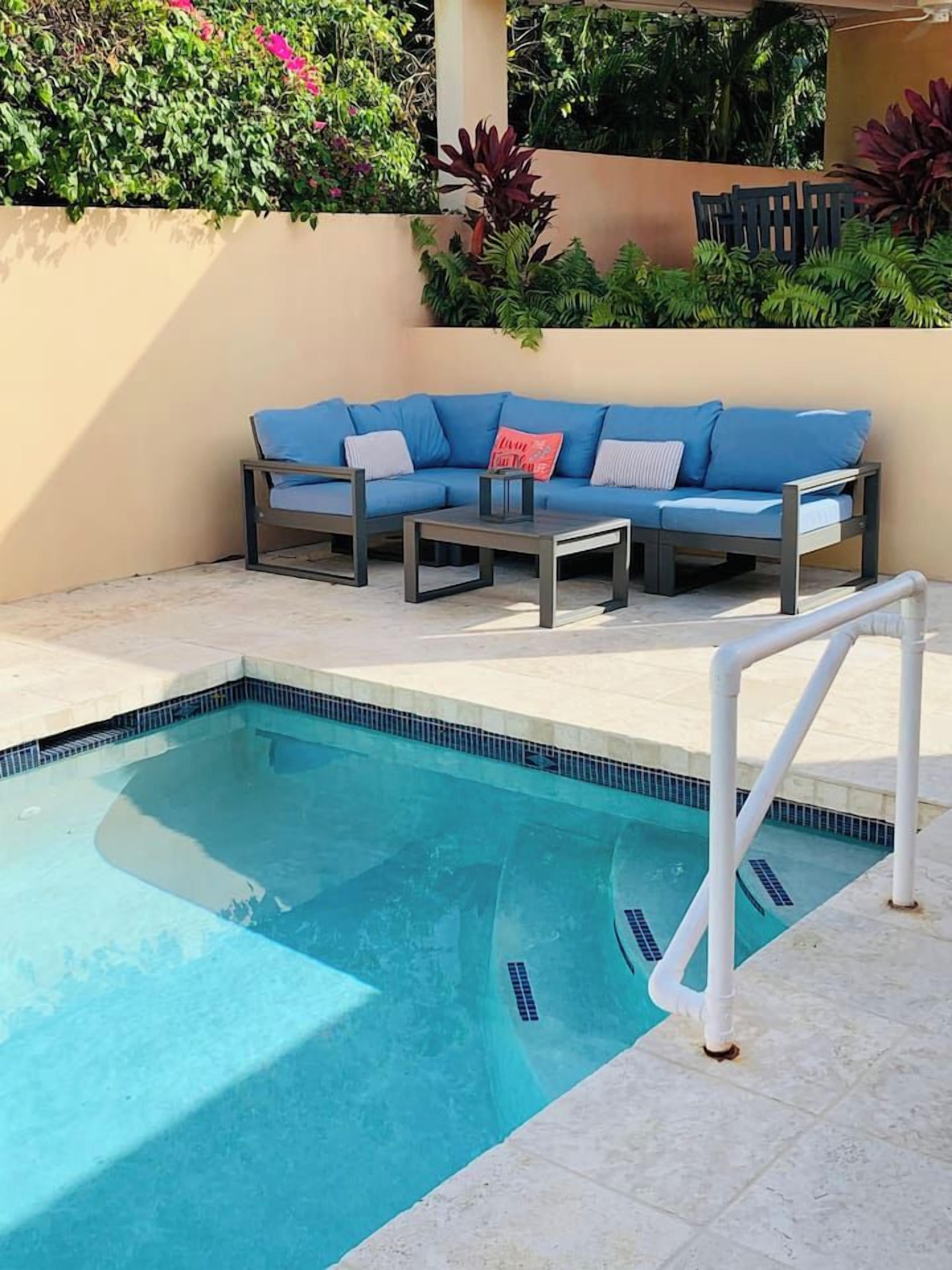 Una piscina con borde de azulejos, una escalera de piscina blanca y un sofá azul con cojines.