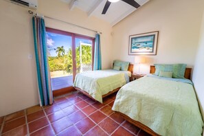 Une chambre avec deux lits, un ventilateur de plafond, un tableau représentant une scène de plage et une vue sur les palmiers à travers la porte vitrée.
