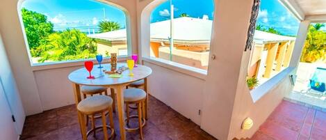 Un pequeño patio con una mesa redonda y cuatro taburetes, dos ventanas que ofrecen vistas a edificios y vegetación, y un suelo de baldosas.