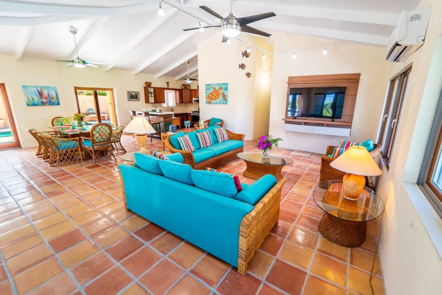 A spacious living area with a large sofa, dining table, and chairs, a kitchen in the background, and a TV mounted on the wall.