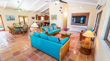 A spacious living area with a large sofa, dining table, and chairs, a kitchen in the background, and a TV mounted on the wall.