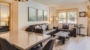 A modern living room with a dining table, sofas, and a large window providing natural light. - Upscale Luxury-Next to Slopes/Golf, View, Hottub (Copper Mountain)