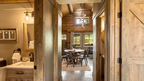 A wooden interior with a bathroom, a dining area with a round table and chairs, and a view of the outdoors through large windows. - Beautiful Log Cabin Near Fishing Access To Yellowstone (Livingston)