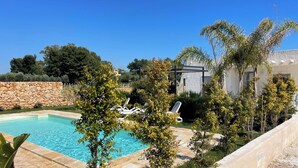 Une piscine entourée de chaises longues, elle-même ceinte d’un mur de pierre et bordée de différents arbres.