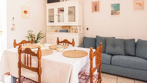 Un coin repas avec une table dressée pour quatre personnes, entourée de chaises en bois. Un meuble avec des portes en verre est adossé au mur, et un canapé est visible au fond.