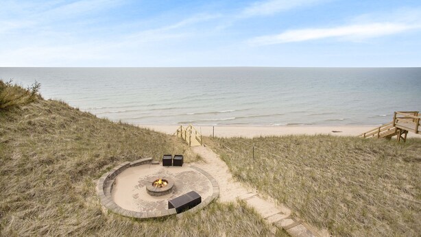 A beachside patio with a fire pit, seating, and a wooden deck leading to the beach. - Custom Lake Michigan Beachfront Vacation Home (Manistee)