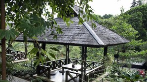 Ein Pavillon mit Holzdach und Geländern, umgeben von üppiger Vegetation und Pflanzen.