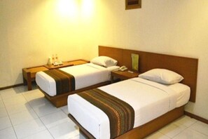Een hotelkamer met twee eenpersoonsbedden, houten hoofdborden, wit beddengoed, een nachtkastje met een lamp en een klein telefoon aan de muur.